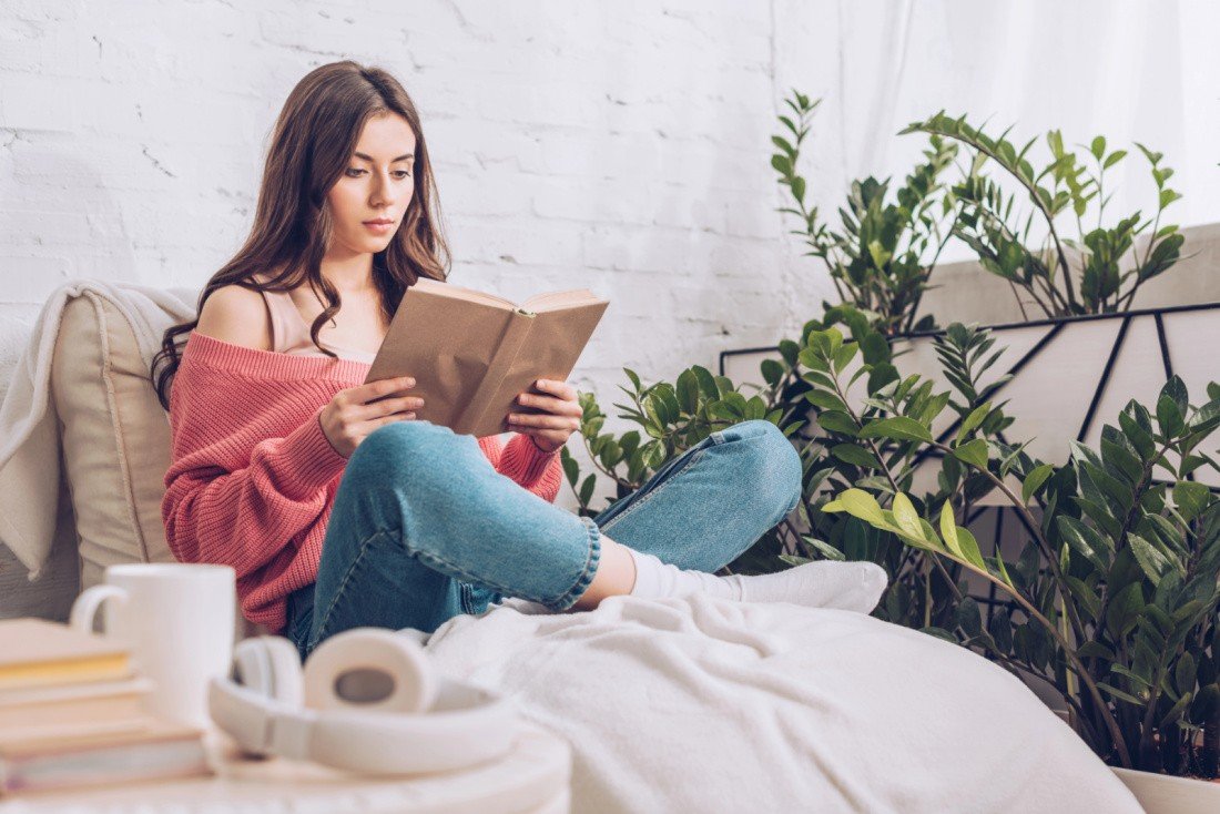 A person wearing a pink sweater and jeans sits on a white sofa, reading a book. Amidst the relaxing home environment, there are plants, a coffee mug, and headphones nearby.