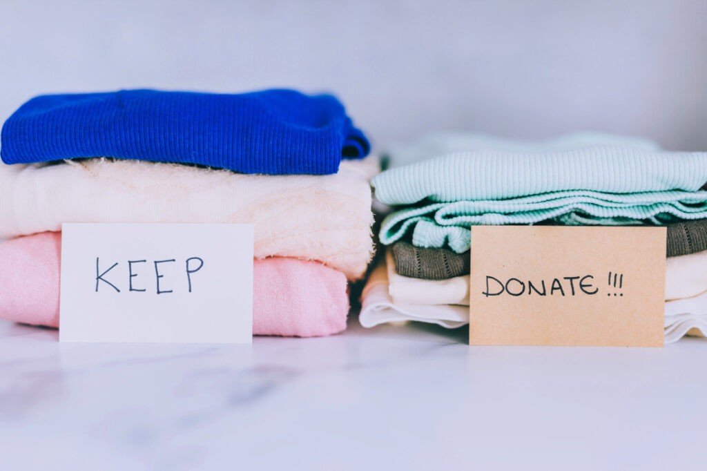 Two piles of folded clothes, one labeled "KEEP" and the other labeled "DONATE!!!", sit on a flat surface in a relaxing home environment.