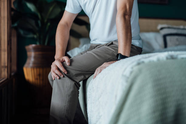 A person sitting on the edge of a bed holds their knee with one hand, wearing a white shirt, khaki pants, and a smartwatch—capturing the struggle with physical symptoms that often go beyond the physical.