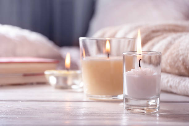 Three lit candles in glass holders rest on a light wooden surface beside folded beige blankets and a closed book—an inviting setup for your evening routine in a softly lit room.