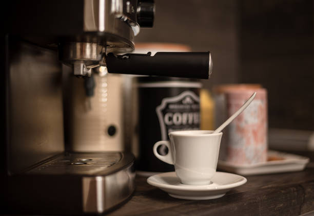 A white espresso cup with a spoon on a saucer sits in front of a coffee machine at a home coffee station, with coffee canisters in the background adding a cozy café vibe.