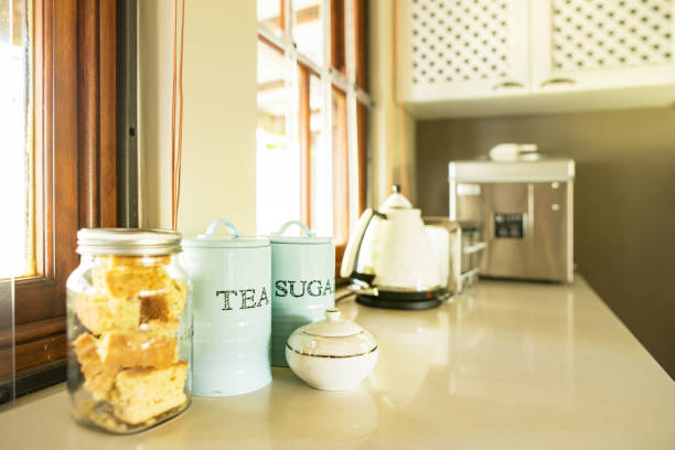 Coffee Station Goals: How to Create a Café Vibe at Home 5 A kitchen countertop with a glass jar of cake, labeled tea and sugar canisters, a sugar bowl, a kettle, and a coffee machine near a window creates a cozy café vibe at home.