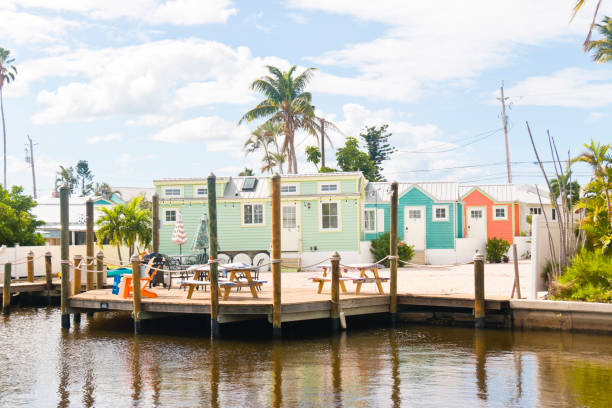Colorful tiny homes line a canal with a wooden dock in the foreground, featuring outdoor seating and palm trees in the background under a partly cloudy sky&mdash;perfect decor tips for making the most of any small space.