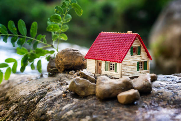 A small model house with a red roof sits on a rock surrounded by stones and a green plant in an outdoor natural setting, perfect inspiration for tiny homes or creative small space decor.