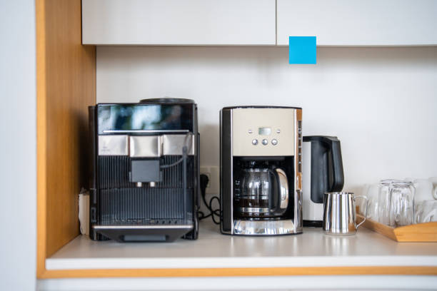 Coffee Station Goals: How to Create a Café Vibe at Home 3 A home coffee station with a coffee machine, drip coffee maker, electric kettle, metal jug, and glass mugs on a countertop beneath a white cabinet brings a cozy café vibe to your kitchen.
