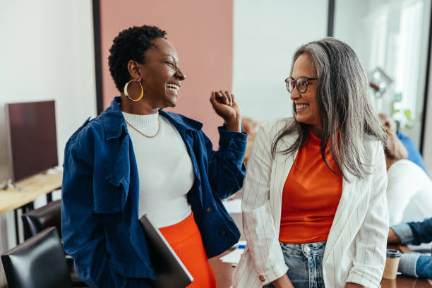 Two women stand indoors, smiling and talking to each other about friendship. One holds a folder. Other people and office furniture are visible in the background, creating a warm atmosphere that supports mental wellness.