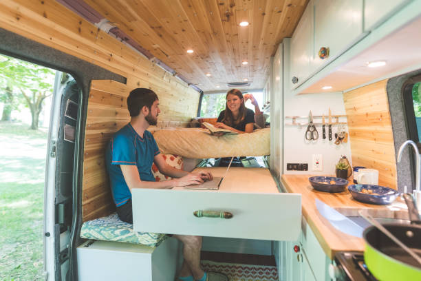 A man uses a laptop at a pull-out table while a woman reads on a bed inside a camper van with wood paneling, compact kitchen, and smart small space decor perfect for fans of tiny homes.