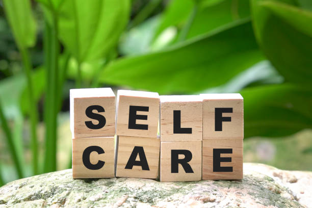 Wooden blocks spelling "SELF CARE" are stacked on a rock with green leaves and plants in the background, highlighting the importance of quick mindfulness even on a busy day.