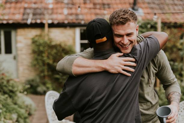 Two men are hugging outdoors in a garden area. One man smiles while holding a cup, while the other, wearing a cap with his back to the camera, shows gentle empowerment through this warm embrace.