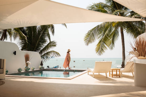 Woman in sunhat stands by the edge of a beachfront pool under palm trees, embracing her travel morning routine with lounge chairs and the ocean in the background—perfect for enjoying travel wellness.