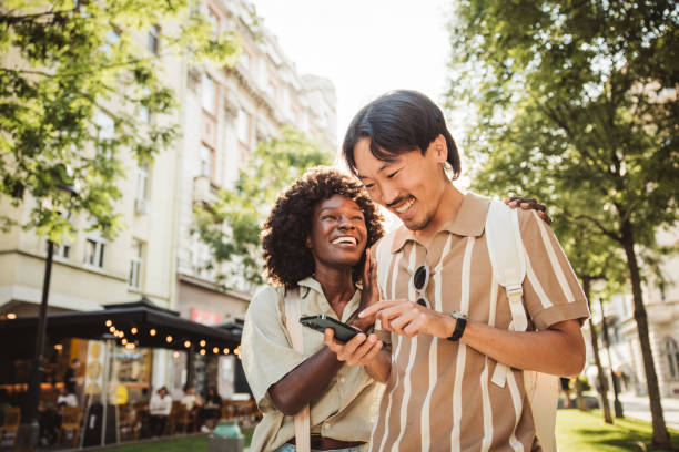 Two people standing outside in a city, smiling and looking at a smartphone together on a sunny day, sharing a moment of empowerment as they gently guide each other through new experiences.