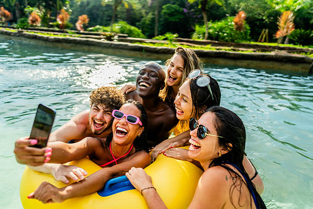 Six young adults in swimsuits sit on a yellow inflatable float in the water, smiling and taking a selfie together. Their laughter captures the empowerment of friendships, with greenery providing a vibrant backdrop.