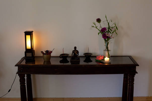 A wooden table against a plain wall holds a lamp, pen holder, Buddha statue, incense holders, a candle, flower vase, and a small decorative item—an inviting Home Self-Care station for moments of peace and reflection.