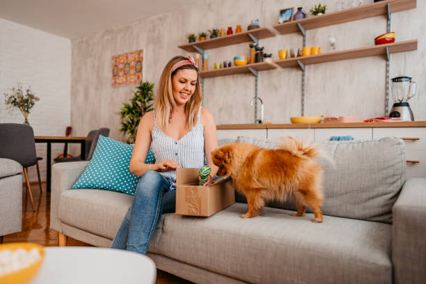 A woman sitting on a couch opens a box while her small brown dog stands beside her, sniffing the contents, creating a pet-friendly sanctuary filled with warmth and mental peace.