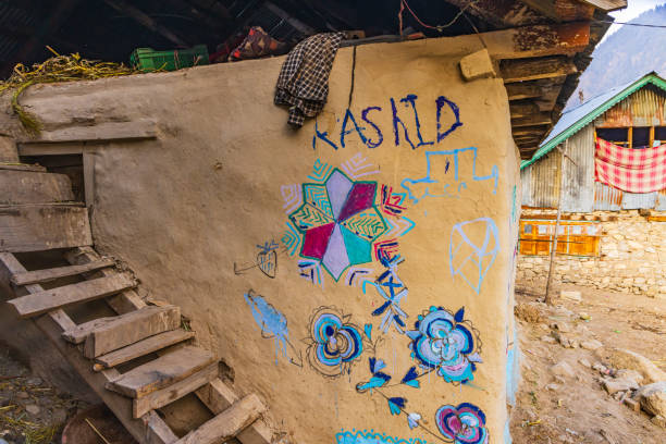 A mud wall with colorful hand-painted designs, serving as a gratitude wall with "KASHID" written on it, stands next to a wooden ladder and traditional houses in the background.