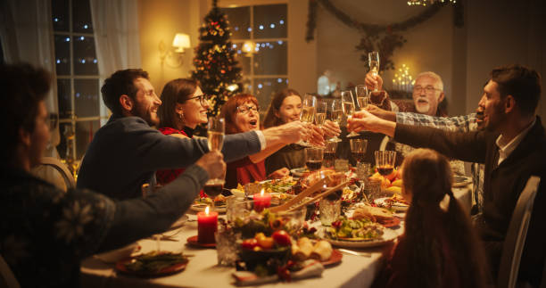 A group of people sitting around a table filled with food raises glasses in a heartfelt toast during a festive holiday dinner, celebrating family connections, with a decorated Christmas tree visible in the background.