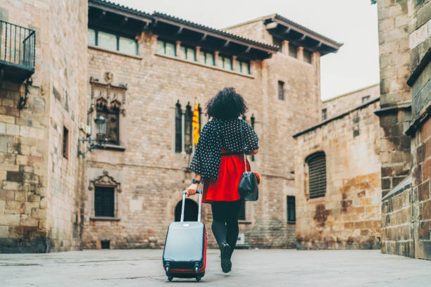 A person practicing self-care, with curly hair, wearing a black top and red skirt, walks on a stone street pulling a suitcase near historic stone buildings&mdash;embracing the spirit of solo travel.