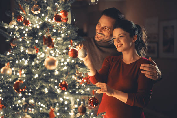 A couple stands by a decorated Christmas tree, smiling and hanging ornaments together in a warmly lit room, embracing heartfelt holiday practices that highlight their family connections.