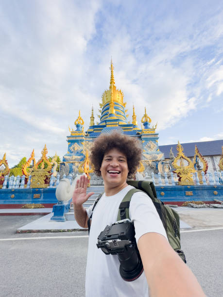 A person with a camera and backpack enjoys solo travel, taking a smiling selfie and waving in front of an ornate blue and gold temple under a partly cloudy sky.