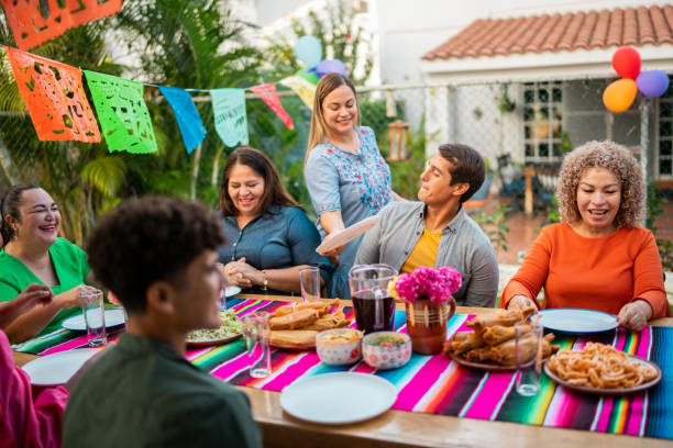 A group of people sit around a table with colorful food and decorations at an outdoor gathering, celebrating heartfelt family connections as they prepare to share a meal.