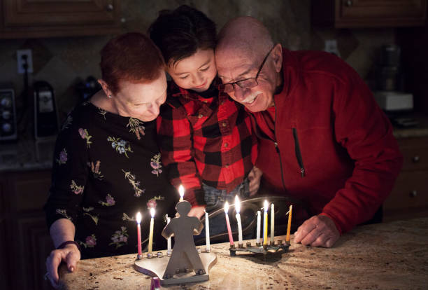 An older woman, a young boy, and an older man share heartfelt smiles while lighting candles on a menorah in the kitchen, embracing family connections and cherished holiday practices during Hanukkah.