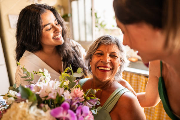 Three women indoors smiling together, with one holding a bouquet of flowers. Sunlight streams in through a window, creating the perfect setting to refresh your space with weekend projects and a touch of home improvement.