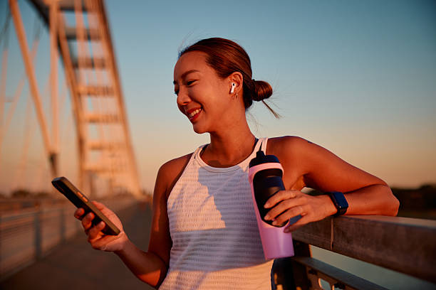 Woman in athletic wear stands on a bridge at sunset, holding a water bottle and looking at her phone with earbuds in, embracing the little joys that make life feel lighter.