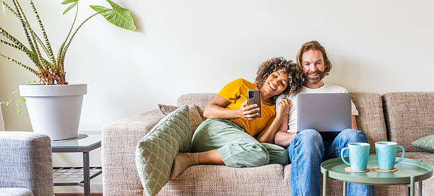 A woman leans on a man while holding a smartphone; the man uses a laptop. They sit on a beige couch with a coffee table in front, savoring the little joys of togetherness, as life feels lighter with a potted plant by their side.