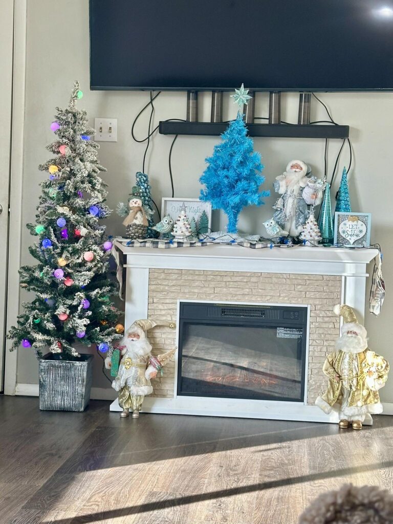 A fireplace decorated with calm, seasonal decor&mdash;white and blue Christmas trees, Santa figurines, and holiday ornaments&mdash;sits beneath a mounted TV.